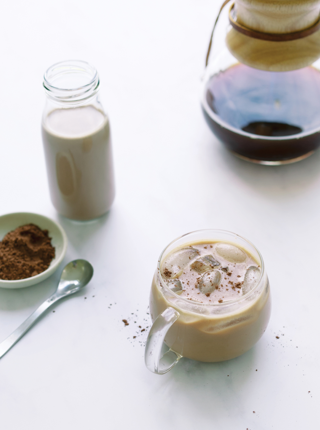 A glass mug filled with coffee and Chocolate Oat Creamer on a counter with a small bowl of ground coffee a silver spoon at the side and a carafe of coffee in the background.