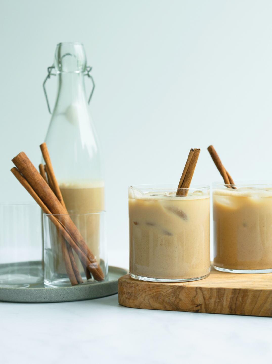 A tray with carafe and glass with cinnamon sticks two glasses of iced Cinnamon Spice Oat Milk Cinnamon on a wooden board.