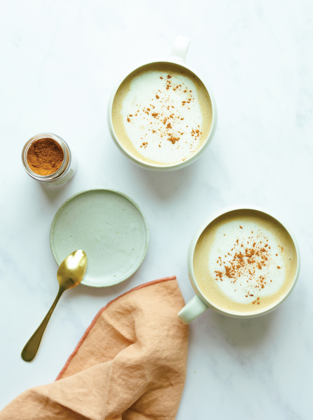 Two large mugs of tea topped with Barista Coconut, Cashew Hemp Milk on a. marble surface with a green plate and light orange napkin and jar of spice.