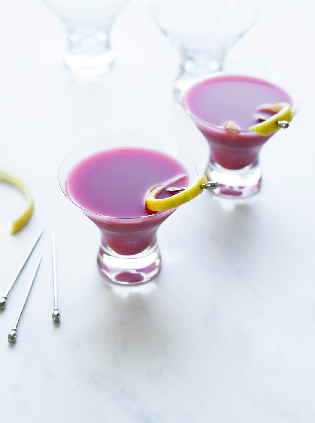 two glasses of dark purple Pom-tini mocktails in low martini glasses with lemon peel and silver skewers