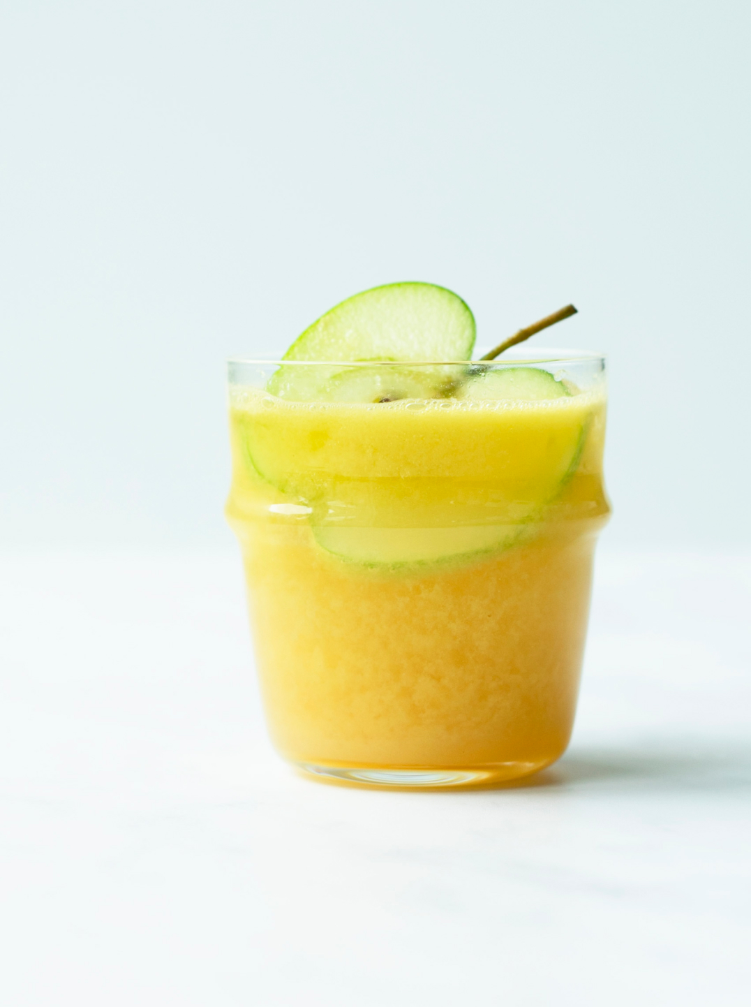 A small glass filled with a deep yellow Apple Sangria juice garnished with a thin slice of green apple.