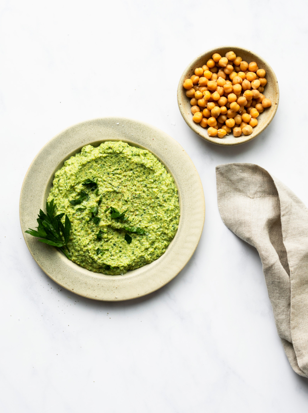 A beautiful green dip in a wide rimmed bowl with linen napkin and small bowl of chic peas