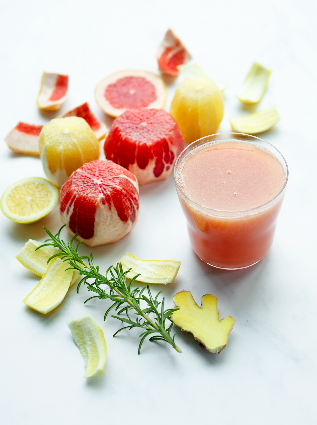 A glass of a beautiful pale pink refreshing rosemary citrus juice surrounded by ingredients pink grapefruit, rosemary and ginger