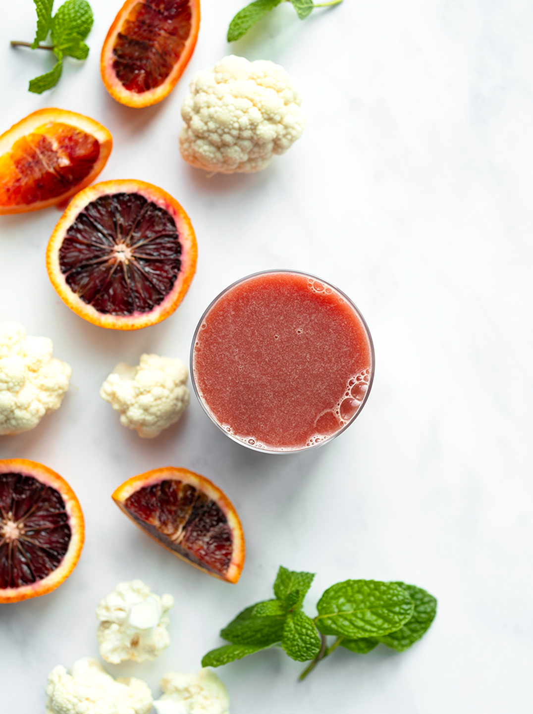 A glass of dark pink minted citrus juice surrounded by sliced blood oranges  cauliflower florets and a sprig of mint.
