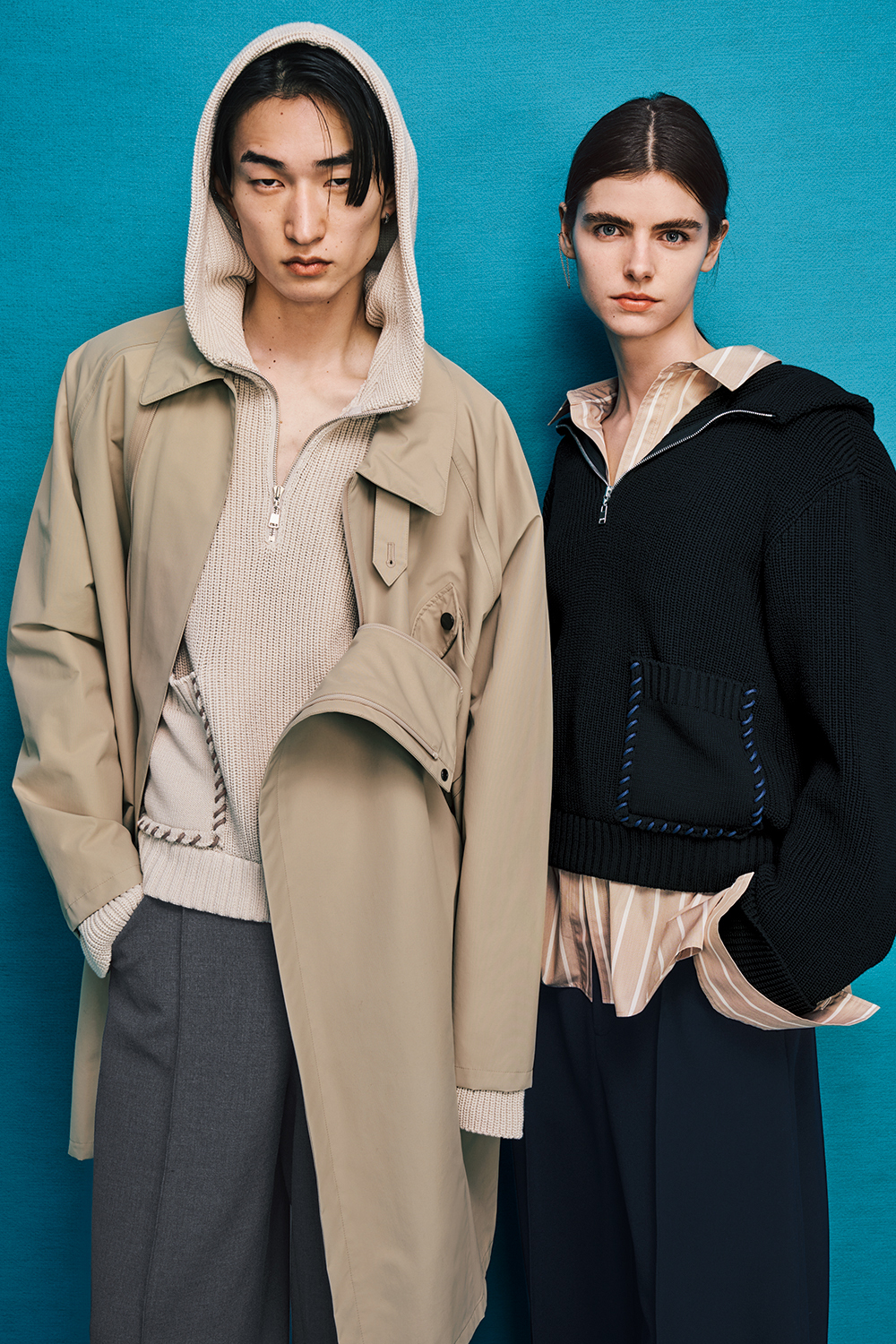 Two models are standing side by side. Model on the left is wearing dark gray pants with a beige coat over a beige top with a hood. Model on the right is wearing black pants with a black jacket over a beige shirt.