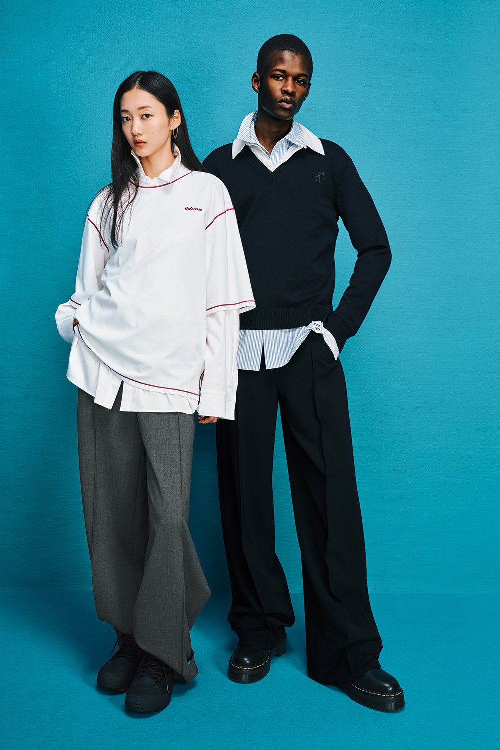 Two models are standing side by side. Model on the left is wearing gray pants with a big white t-shirt over a white shirt. Model on the right is wearing black pants with a black sweater over a striped shirt.