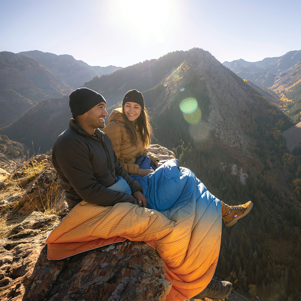 Two people enjoying the view atop a mountain with a Rumpl Original Puffy Blanket spread out over their legs to keep cozy.