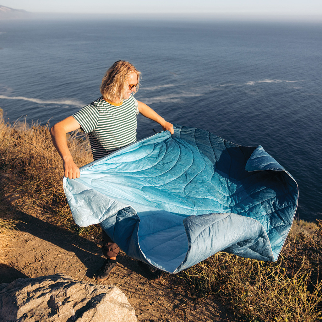 A person dusting off their Rumpl Original Puffy Blanket while on the trail.