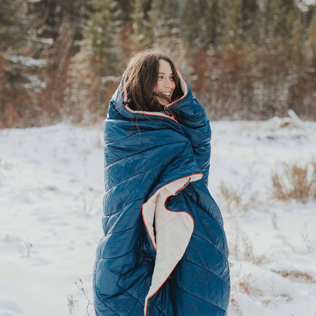 A woman walking out in the snow while wrapped head to toe in a Rumpl Sherpa Puffy Blanket.