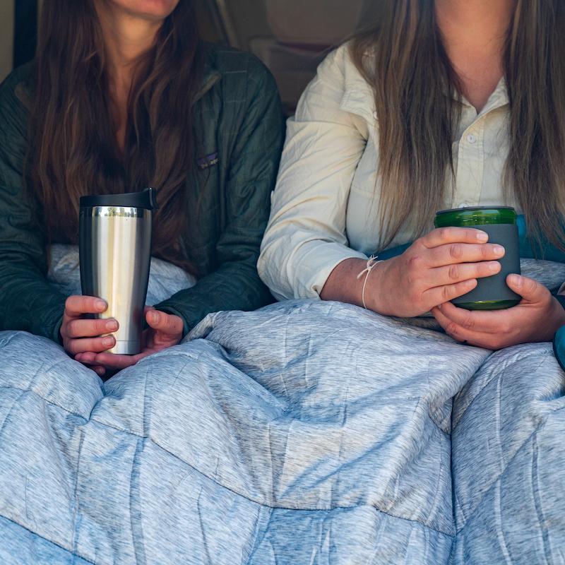 two people sitting shoulder-to-shoulder at the back of a camper van, holding drinks and warming up with the 2-person sized Rumpl Original Puffy Blanket