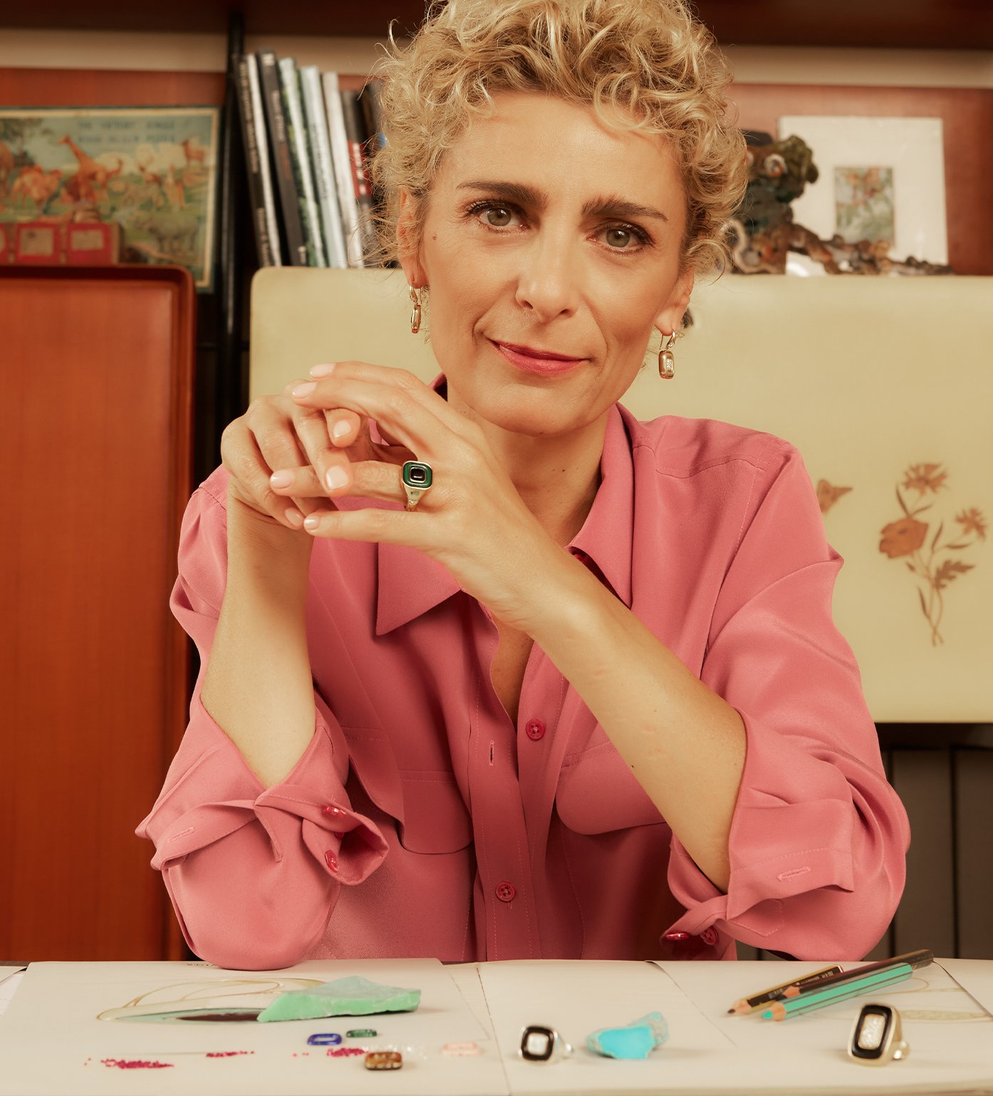 Portrait of Francesca Villa. She has short, curly blond hair and is wearing a pink collared shirt. On her desk are two of her ring peices. 