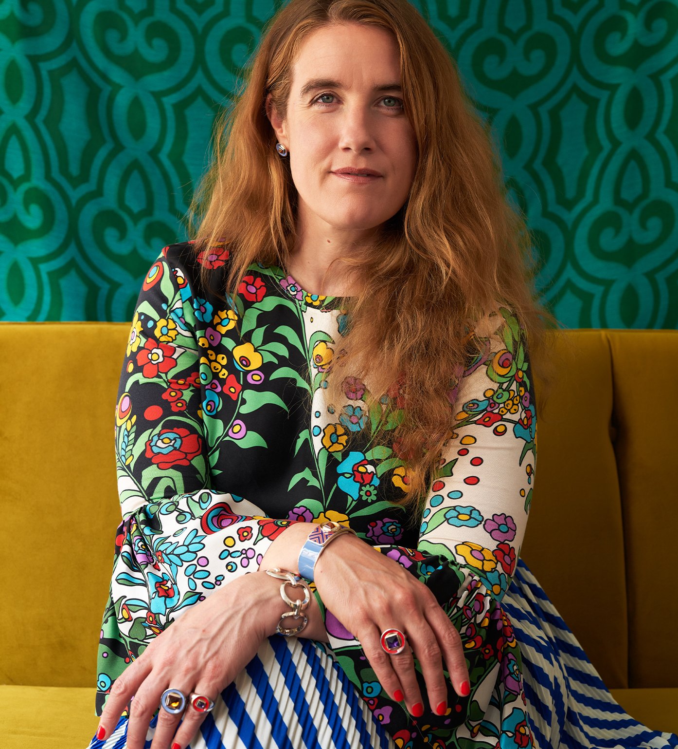 Portrait of Alice Cicolini. She has light eyes, fair skin, and long reddish hair. She is wearing a colorful dress with a 70s-era floral pattern, and is wearing several of her Mad Mod jewely peices. 