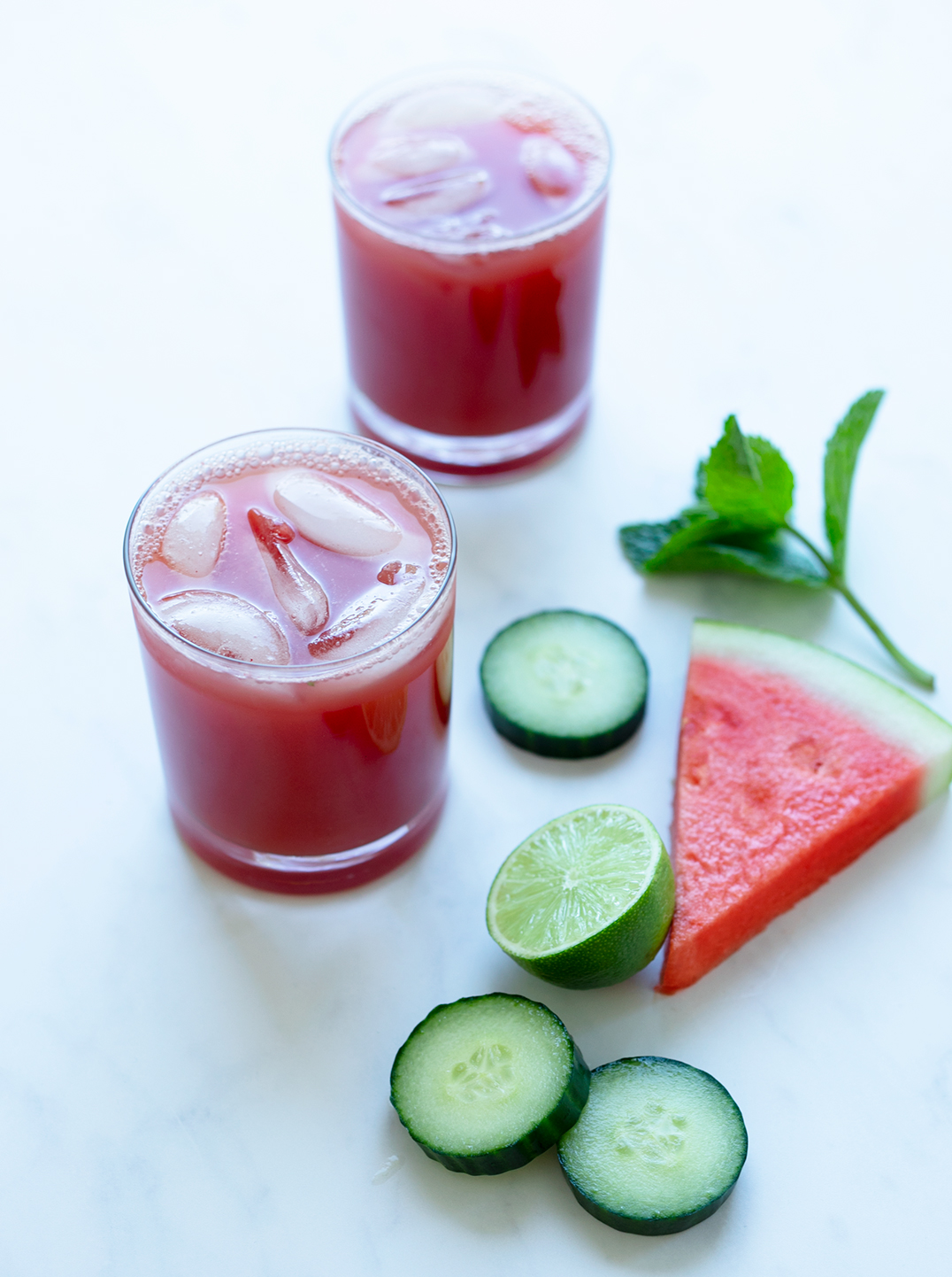 a glass of Watermelon Refresher juice with ice, surrounded by slices of cucumber and a wedge of watermelon

