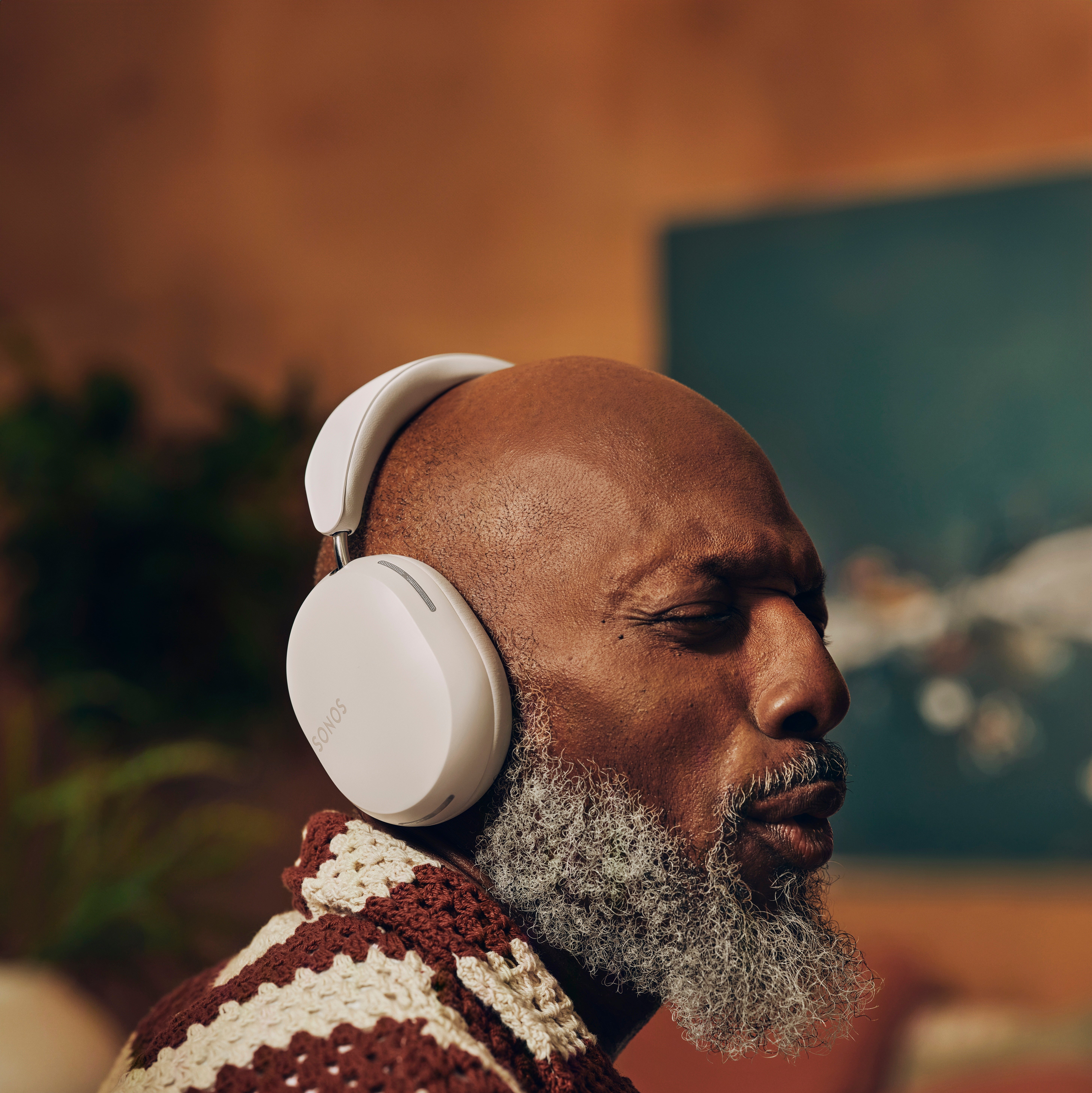 Man with gray beard wearing white wireless headphones, eyes closed, enjoying music in a cozy living room.