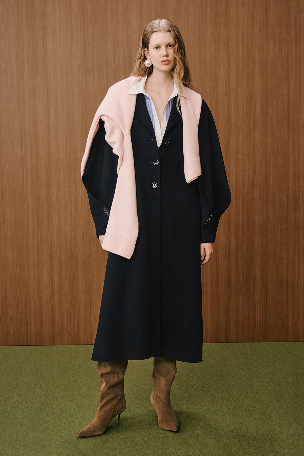 A model is wearing a long black coat layered over a white shirt and striped blouse, styled with a soft pink knit draped over the shoulders and brown knee-high boots.