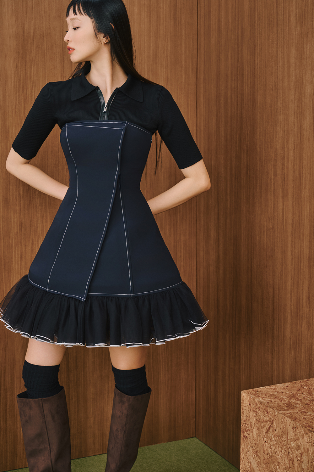 A model is wearing a navy fitted dress with contrast stitching and a sheer ruffled hem, paired with black socks and brown knee-high boots.