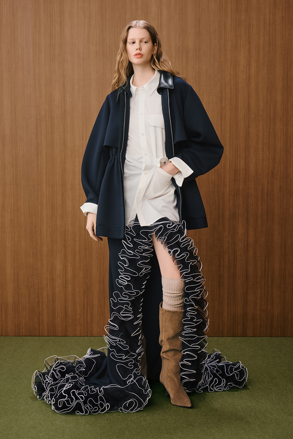A model is wearing a white button-up shirt layered under a navy oversized jacket, paired with black pants featuring dramatic ruffled detailing and brown knee-high boots.
