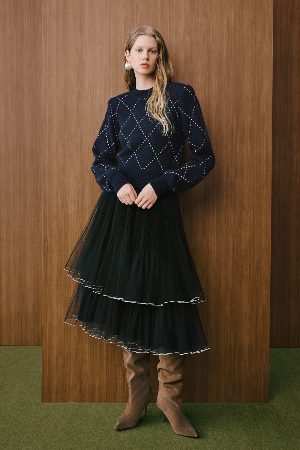 A model is wearing a navy argyle-patterned knit sweater paired with a layered black tulle midi skirt and brown knee-high boots.