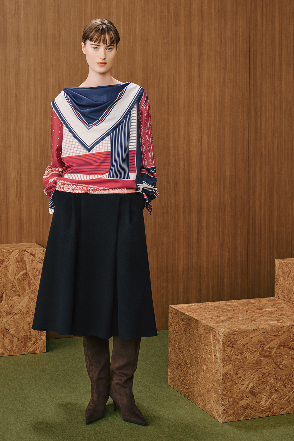 A model is wearing a red, navy, and white patterned long-sleeve top paired with a black midi skirt and brown knee-high boots.