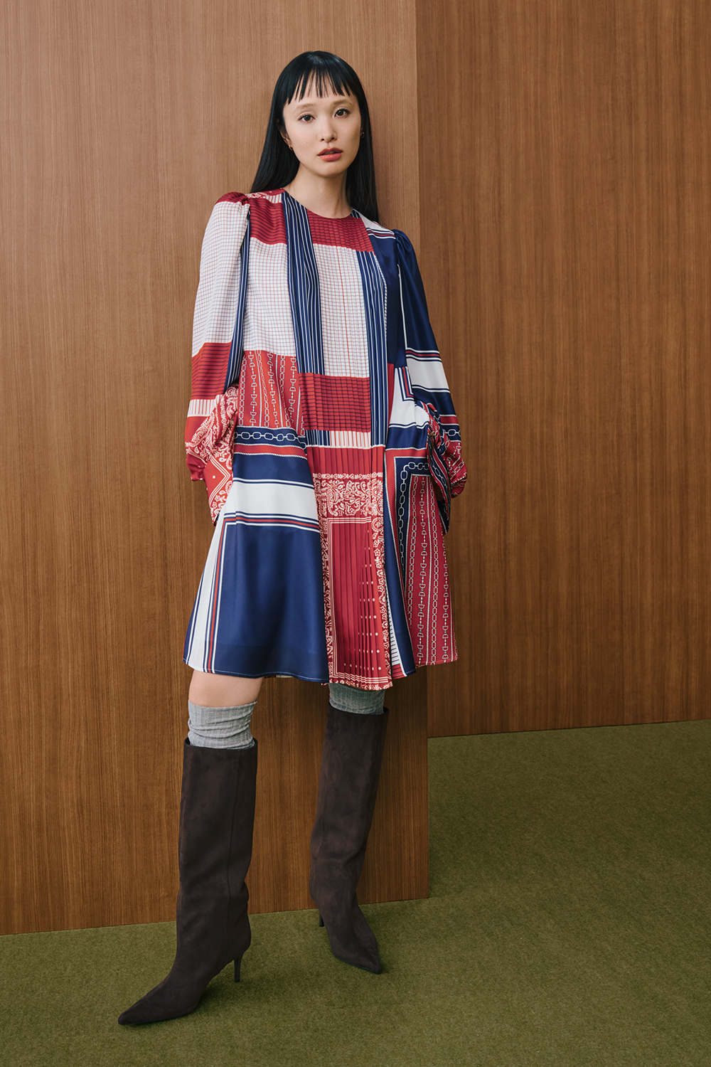 A model is wearing a red, navy, and white patchwork-patterned long-sleeve dress paired with brown knee-high boots.
