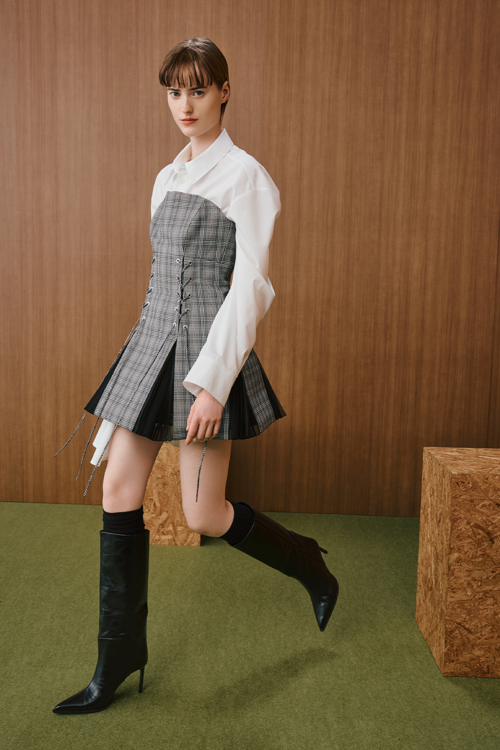 A model is wearing a white shirt layered under a gray plaid corset-style mini dress with pleated detailing, paired with black knee-high boots.