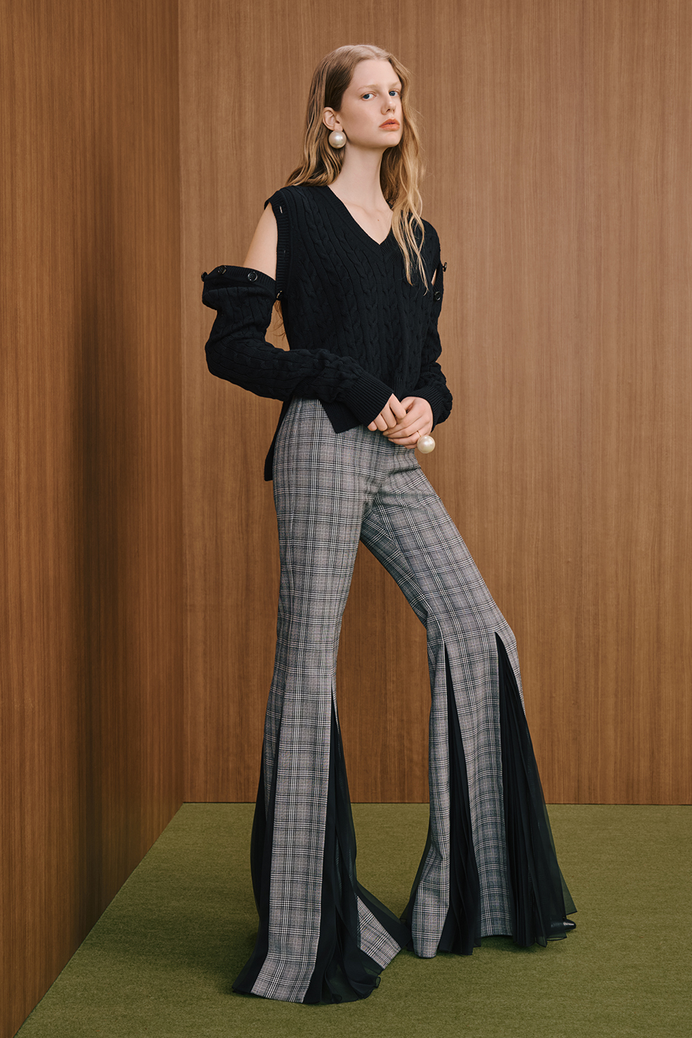 A model is wearing a black sleeveless knit top with shoulder cutouts, paired with gray plaid flared pants featuring sheer panel details.