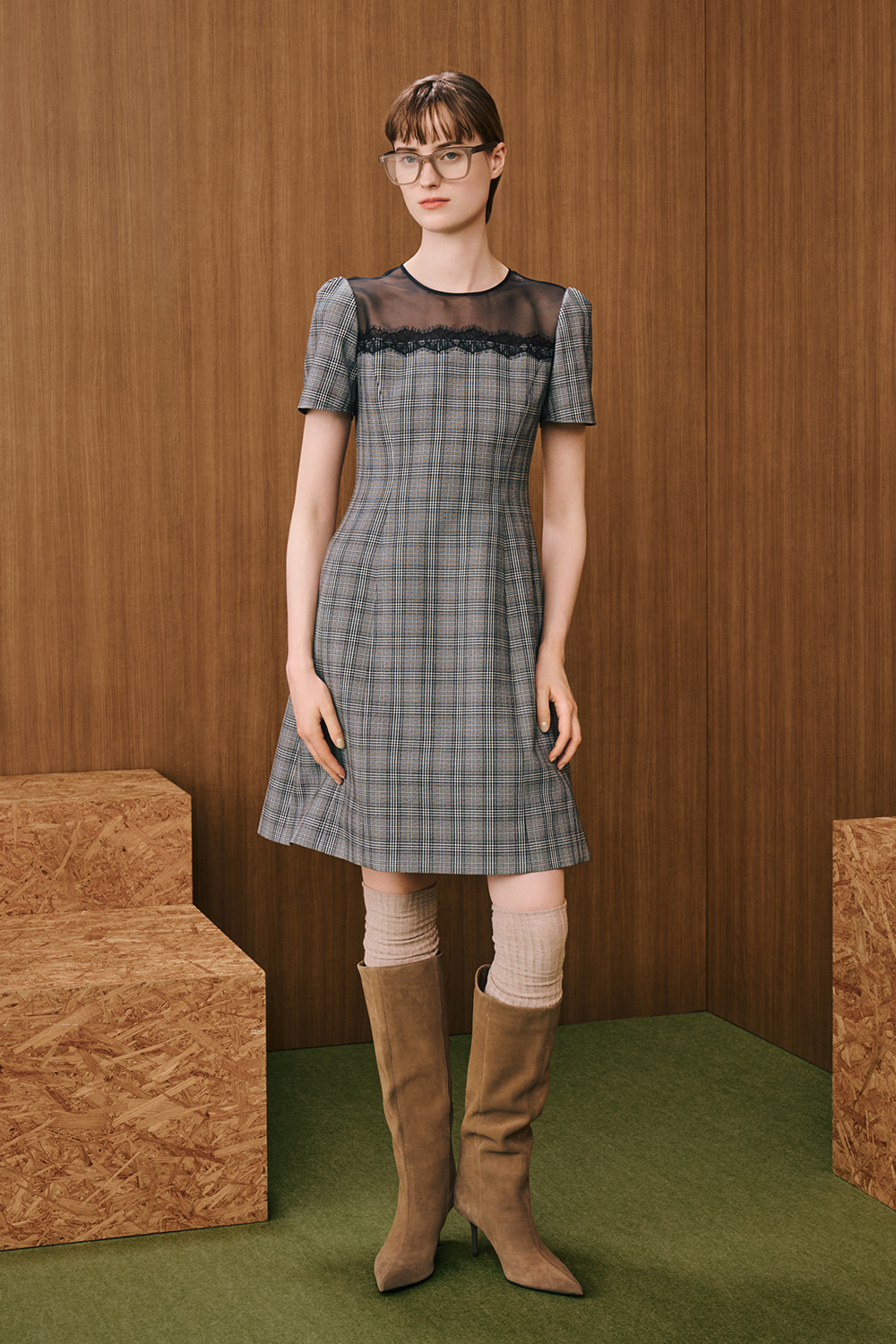 A model is wearing a gray plaid short-sleeve dress with a sheer black yoke detail, styled look with brown knee-high boots.