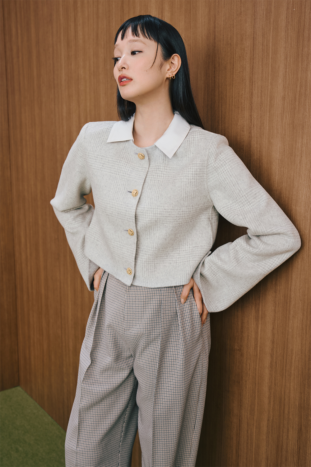 A model is wearing a light gray cropped jacket with gold buttons layered over a white collared top, paired with gray check-patterned wide-leg pants.