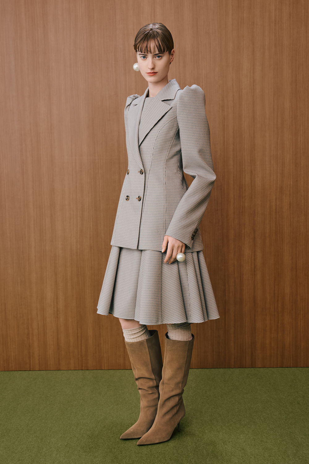 A model is wearing a gray check-patterned tailored jacket with structured shoulders, paired with a matching pleated skirt and brown knee-high boots.
