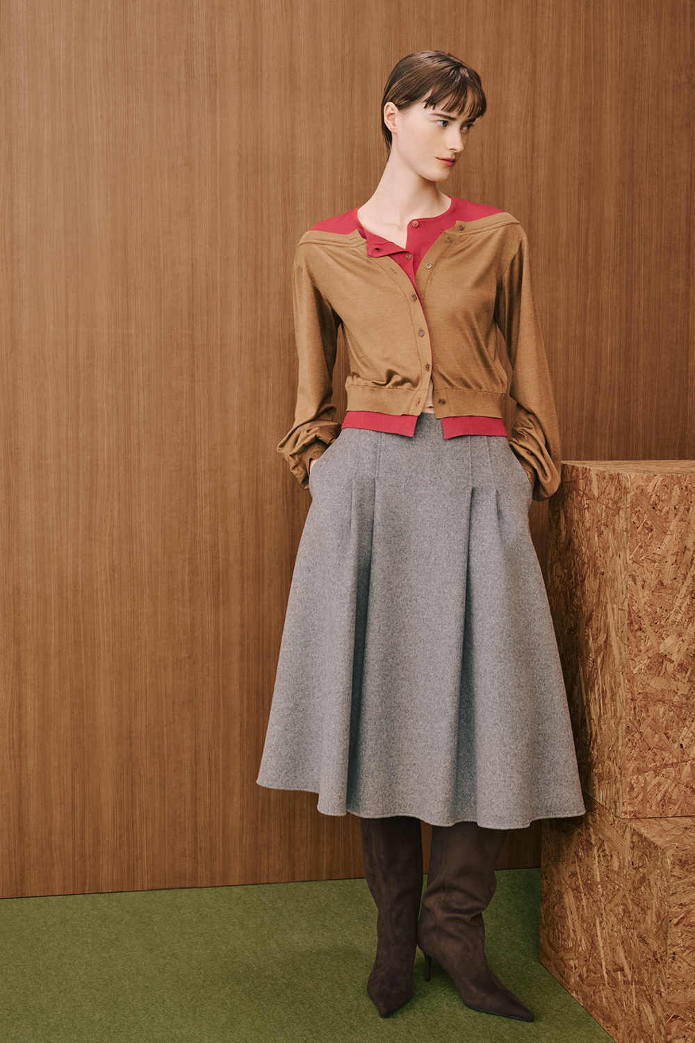 A model is wearing a camel cardigan layered over a red top, paired with a gray flared midi skirt and brown knee-high boots.