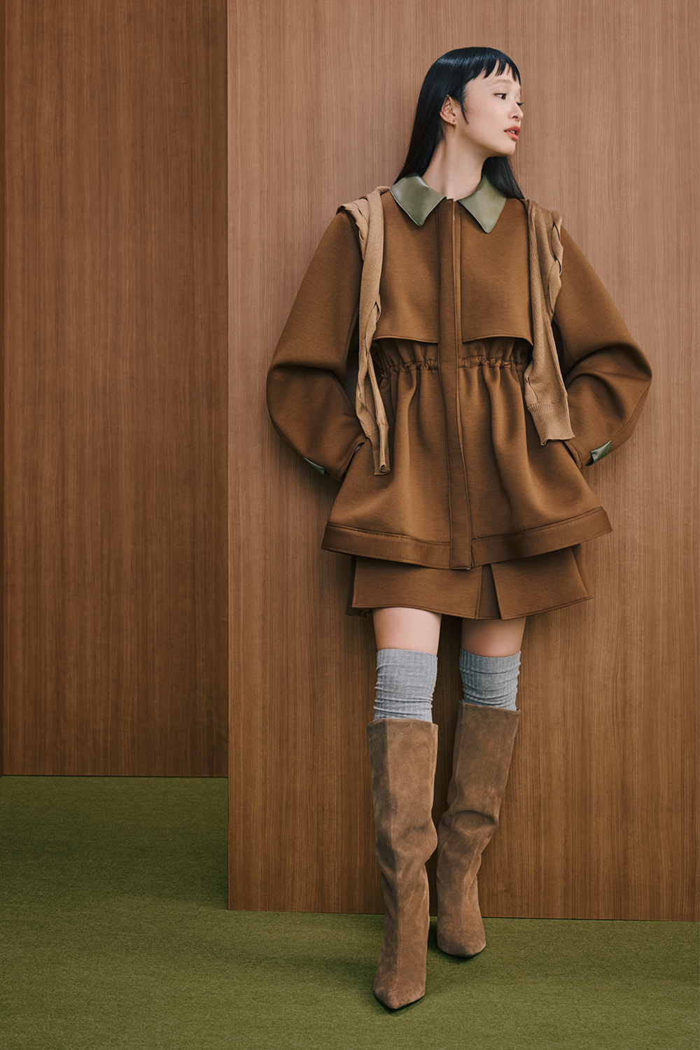 A model is wearing a brown hooded dress with a gathered waist and leather collar detail, paired with brown knee-high boots.