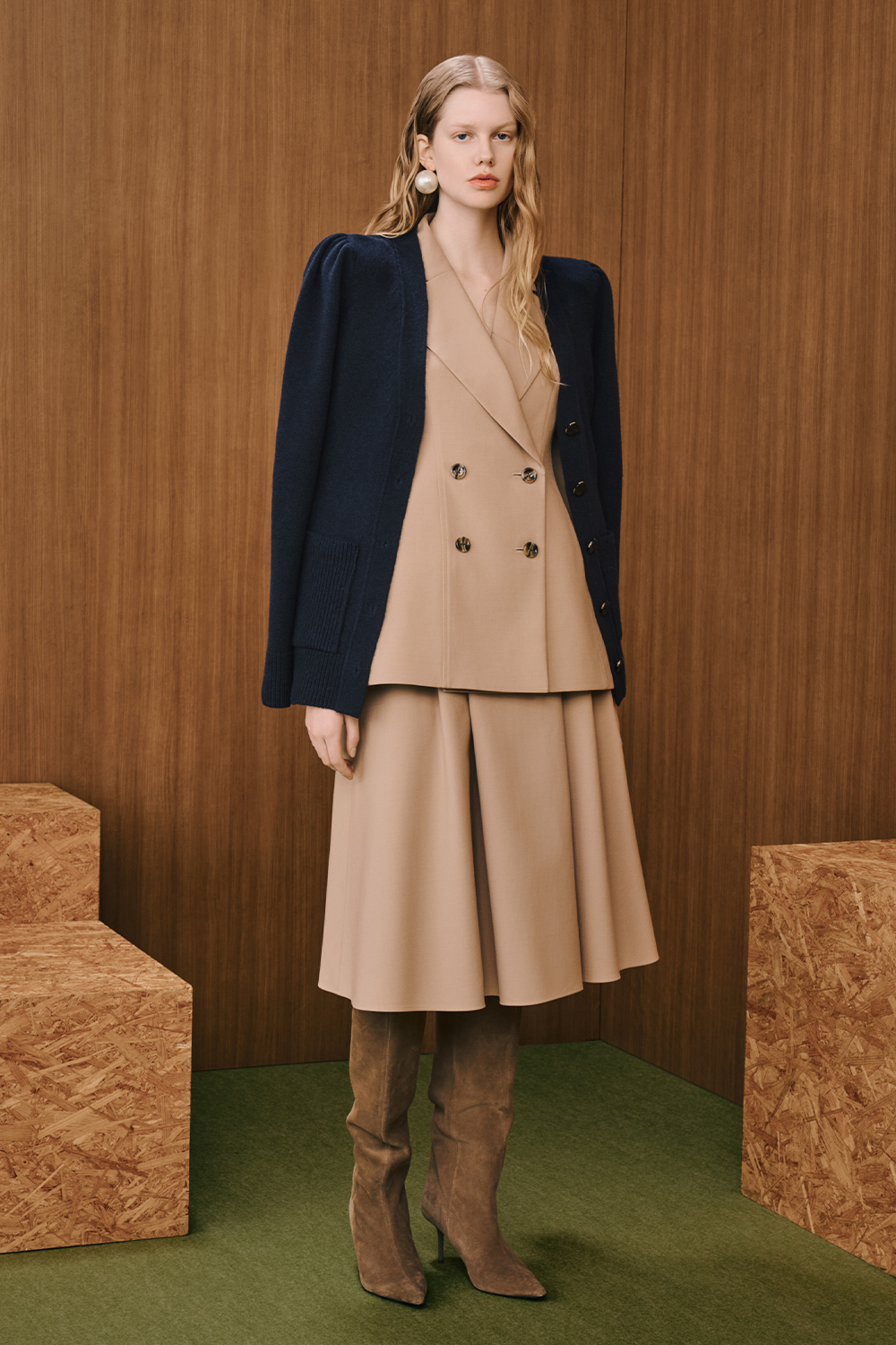 A model is wearing a beige double-breasted vest layered under a navy cardigan, paired with a beige flared midi skirt and brown knee-high boots.