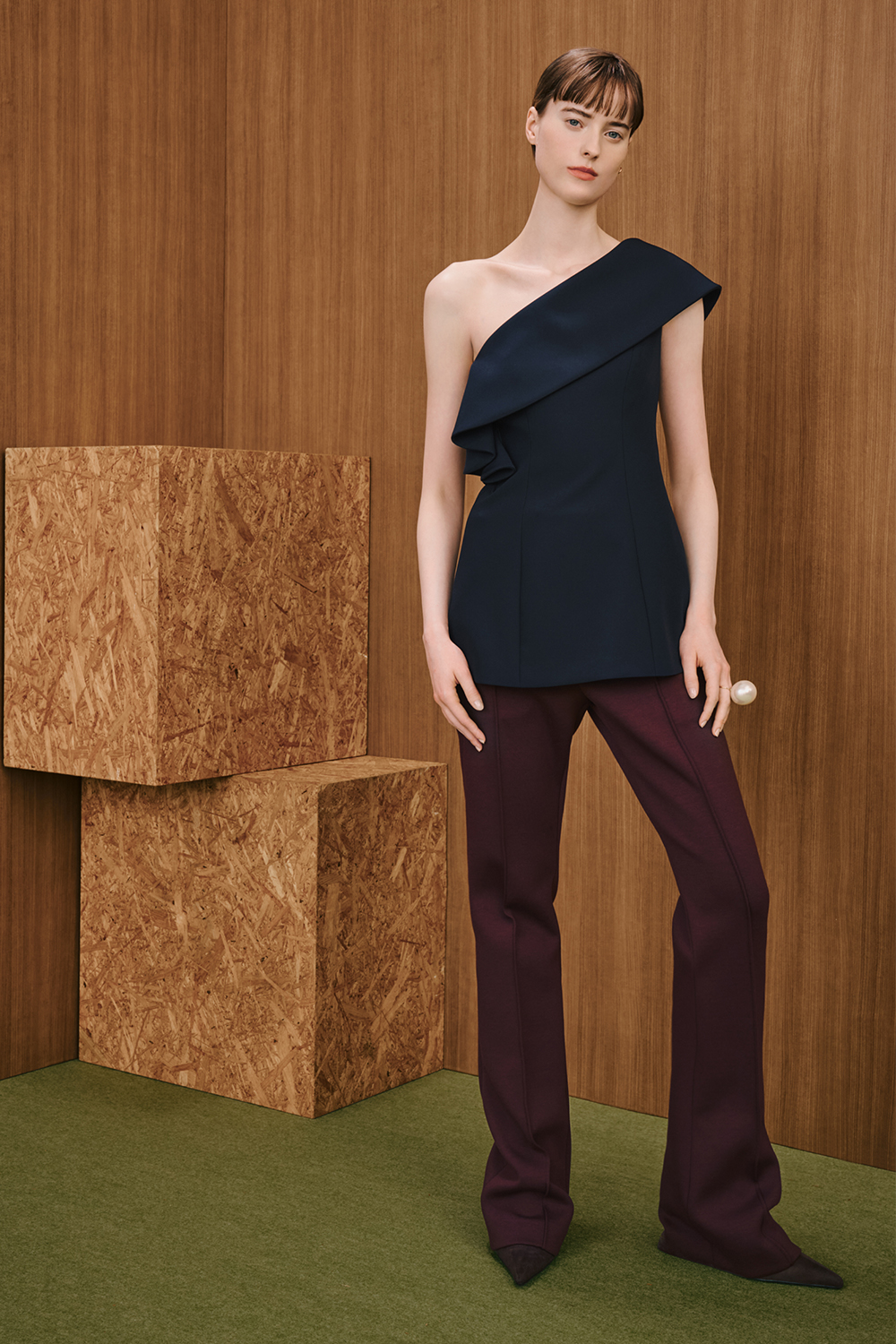 A model is wearing a black one-shoulder top with an asymmetric neckline, paired with burgundy flared trousers.