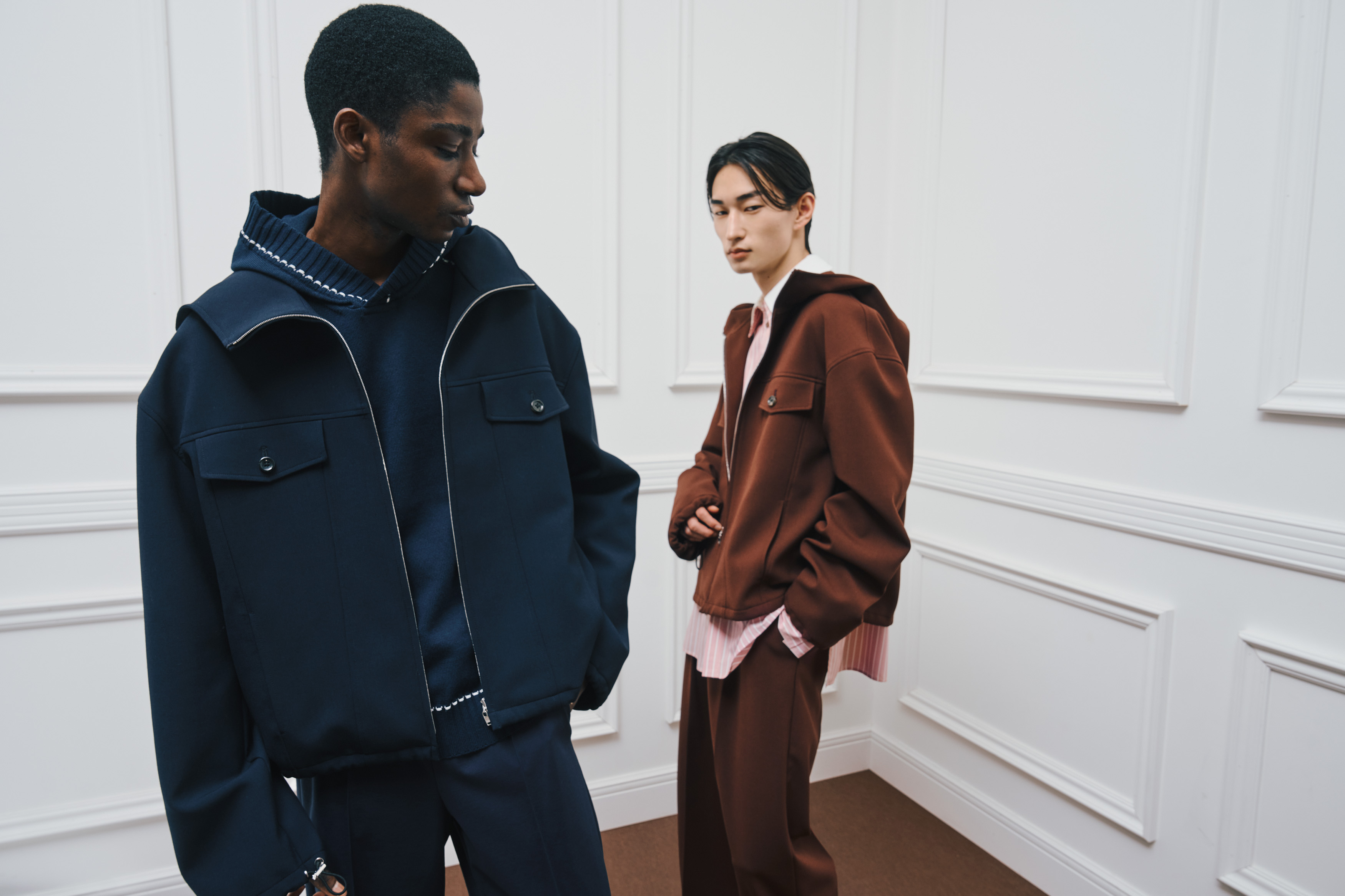 Two models are standing together. The model on the left is wearing a navy hooded jacket with flap pockets, paired with navy wide-leg pants. The model on the right is wearing a brown zip-up jacket layered over a pink striped shirt, paired with brown wide-leg pants.