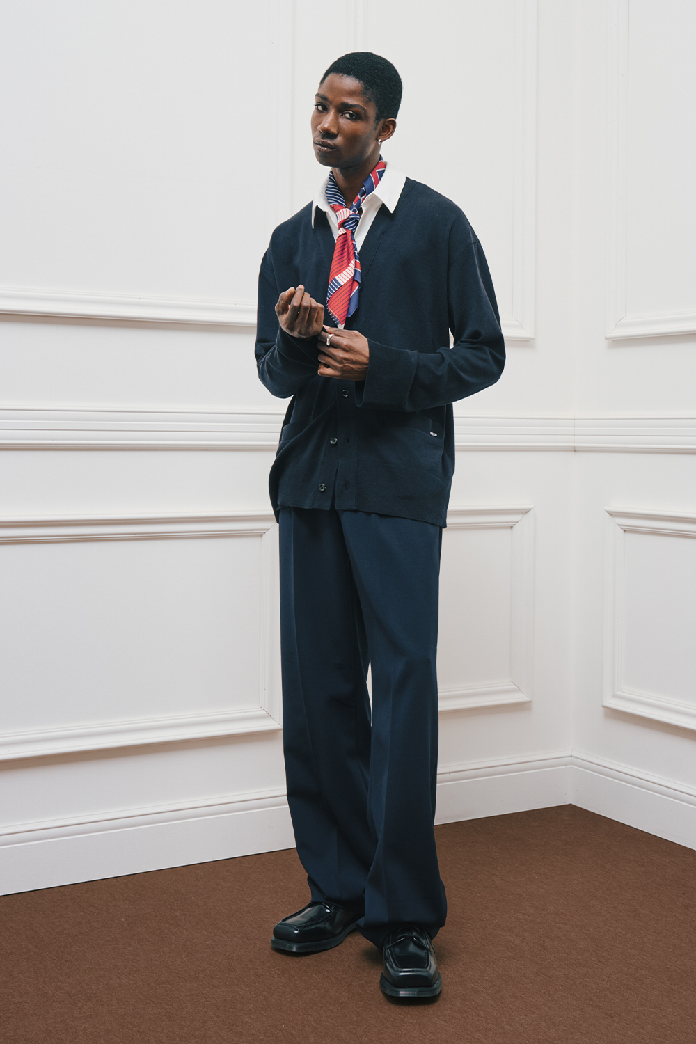 A model is wearing a navy cardigan layered over a white shirt, styled with a red, navy, and white striped scarf and navy wide-leg pants.