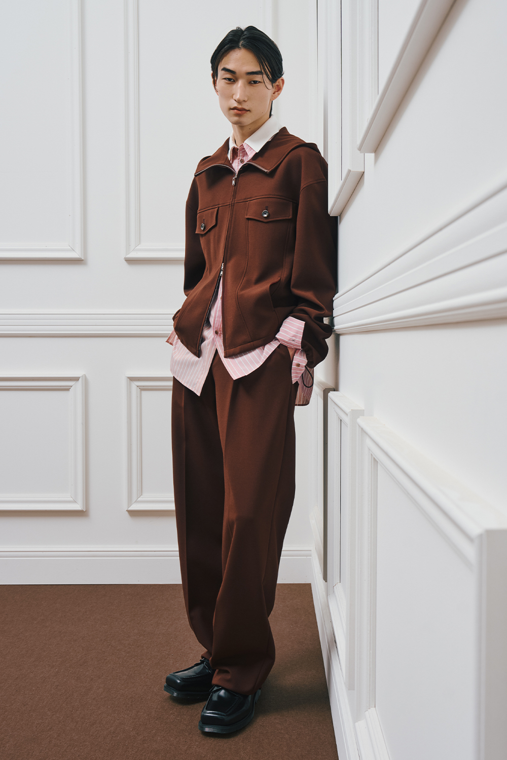 A model is wearing a brown zip-up jacket layered over a pink striped shirt, paired with brown wide-leg pants and black shoes.