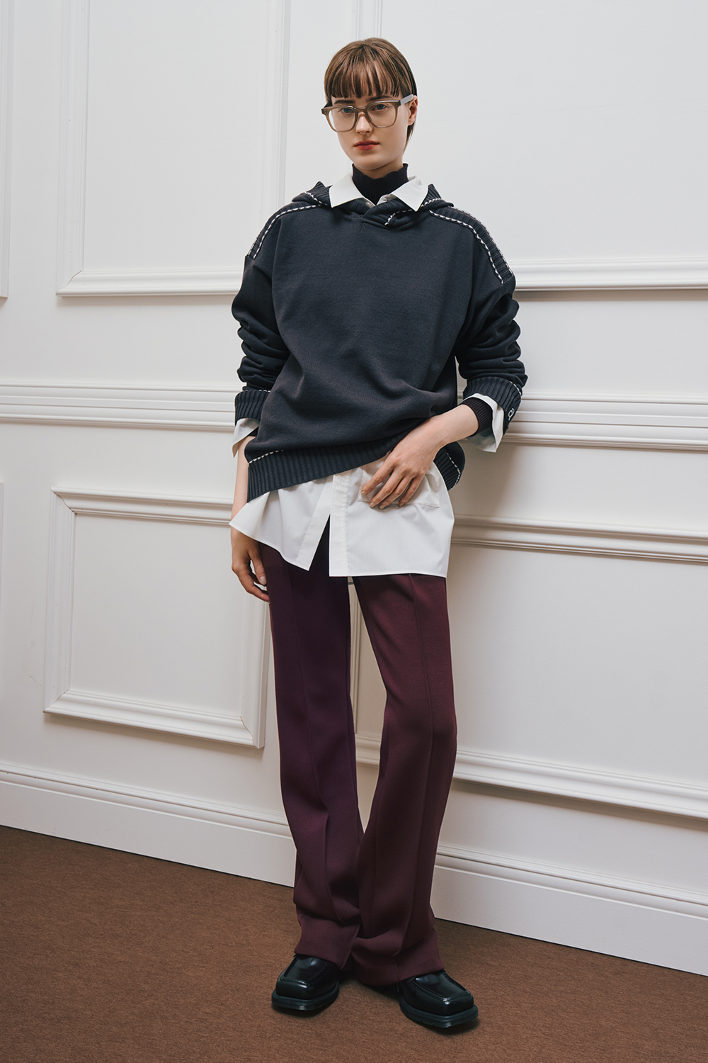 A model is wearing a dark navy knit sweater layered over a white shirt, paired with burgundy straight-leg pants and black shoes.