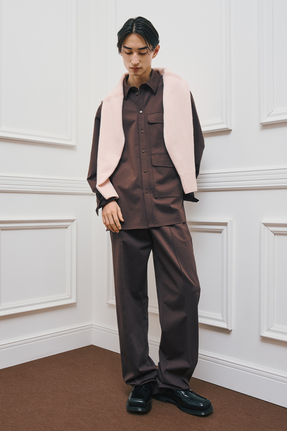 A model is wearing a brown button-up shirt layered under a soft pink knit draped over the shoulders, paired with brown wide-leg pants and black shoes.