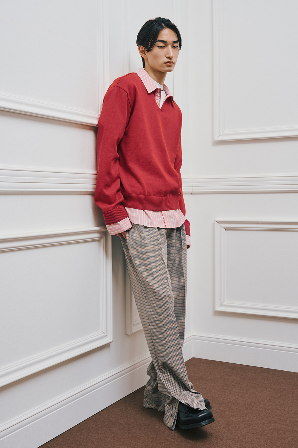 A model is wearing a red knit sweater layered over a pink striped shirt, paired with gray check-patterned wide-leg pants and black shoes.