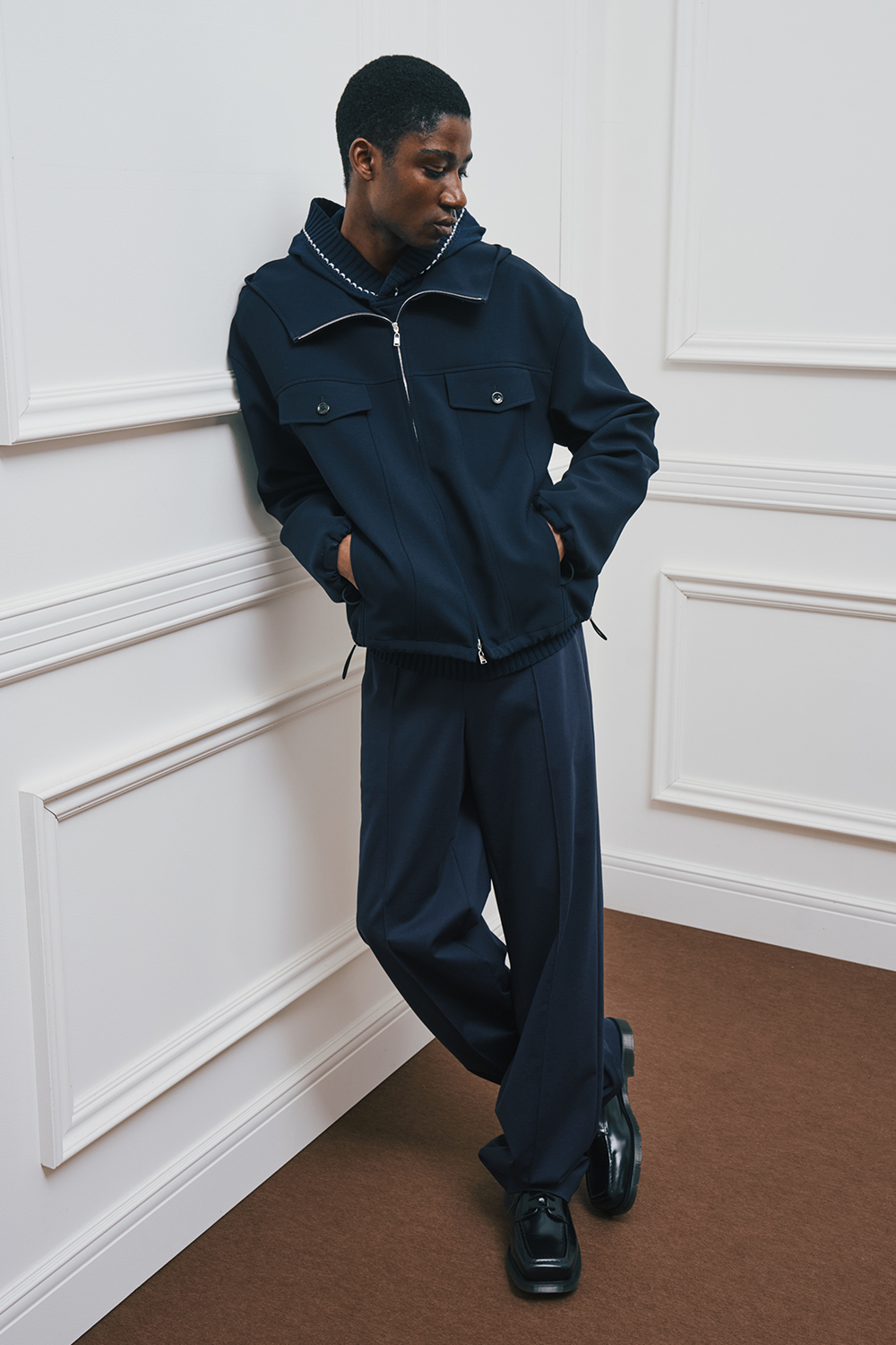 A model is wearing a navy hooded jacket with flap pockets, paired with navy wide-leg pants and black shoes.