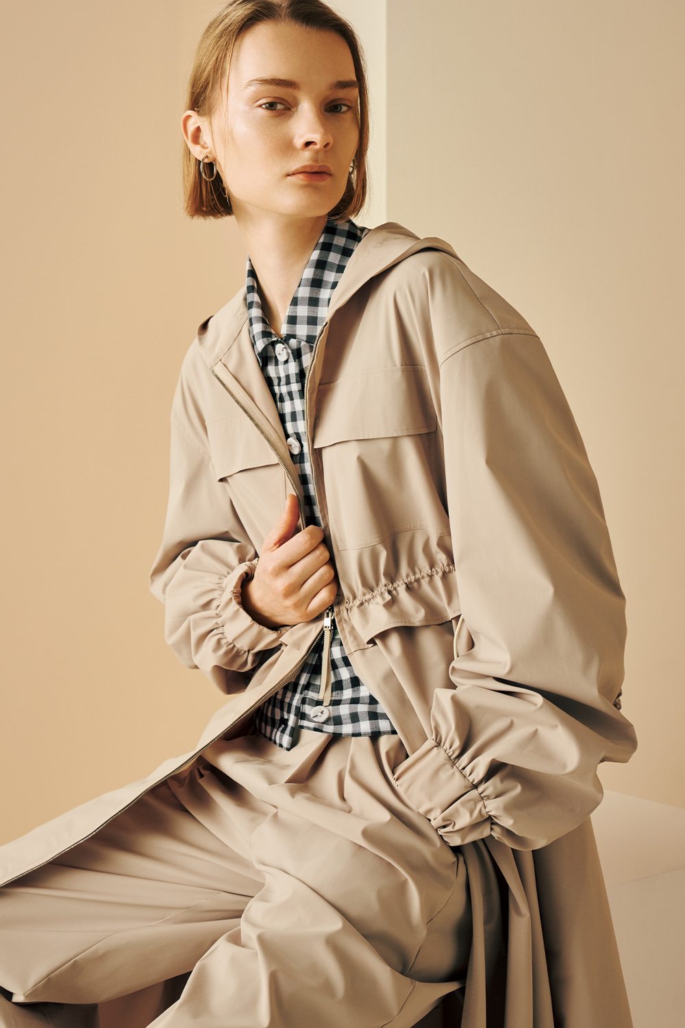 Model is wearing a beige hooded coat over a black-and-white gingham shirt, paired with matching beige pants.