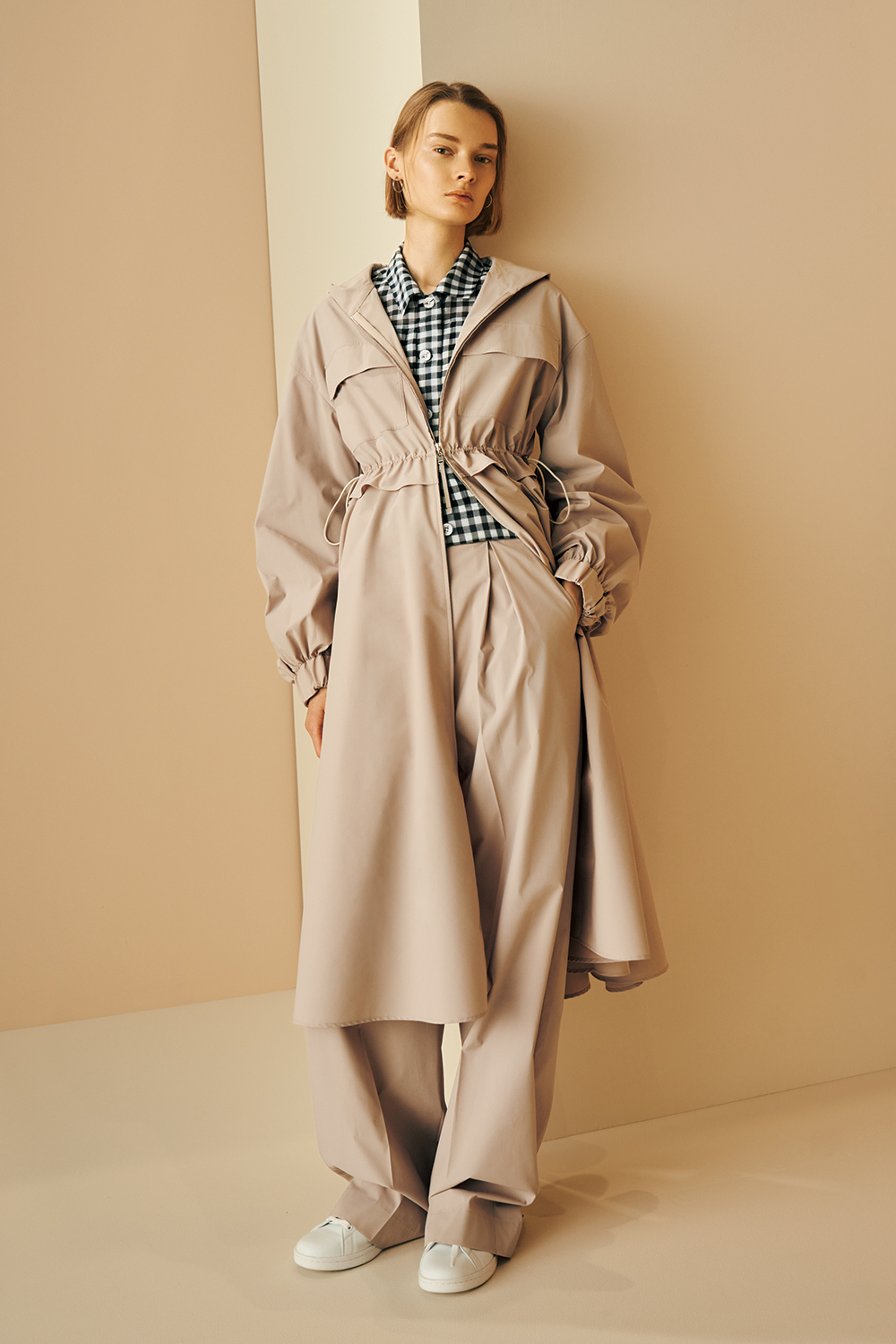 Model is wearing a beige hooded long coat with a cinched waist, layered over a black-and-white gingham shirt, paired with wide-leg beige pants and white sneakers.