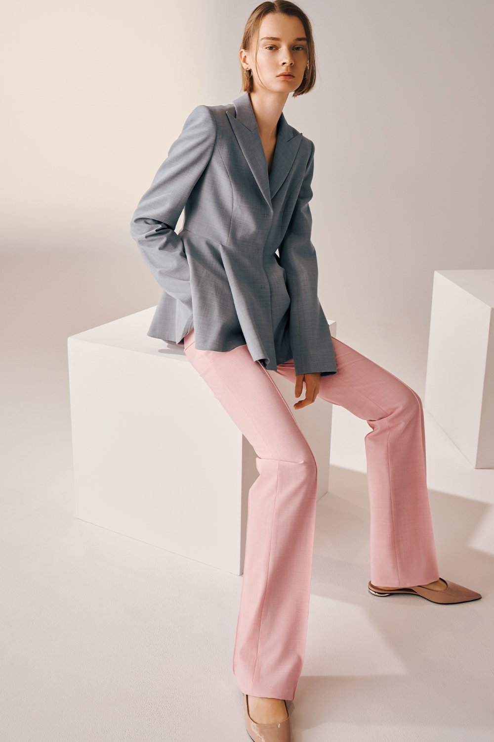 Model is wearing a tailored light grey blazer, pink flared trousers, and beige pointed-toe flats.