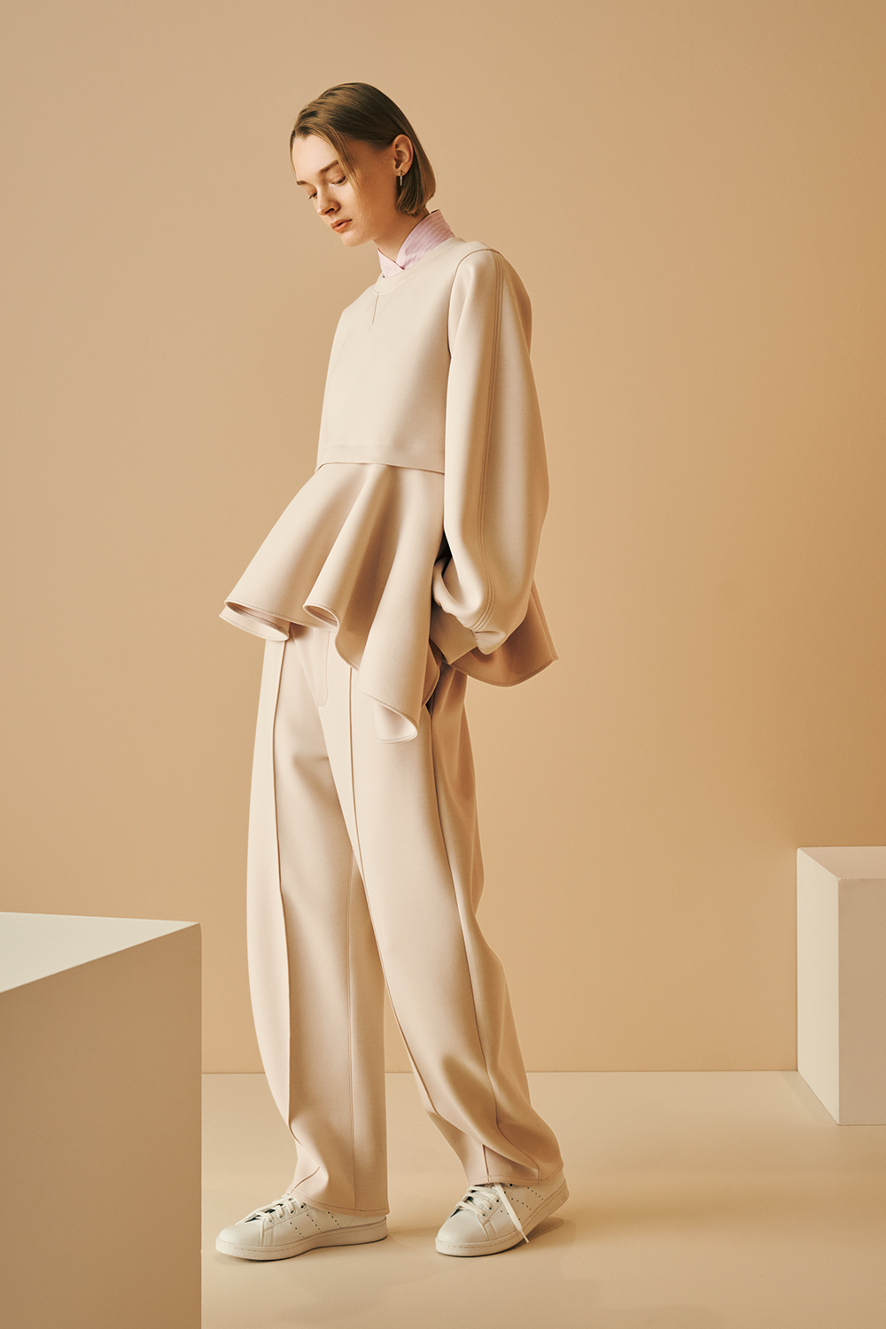 Model is wearing a beige oversized peplum top with voluminous sleeves, wide-leg matching pants, and white sneakers.