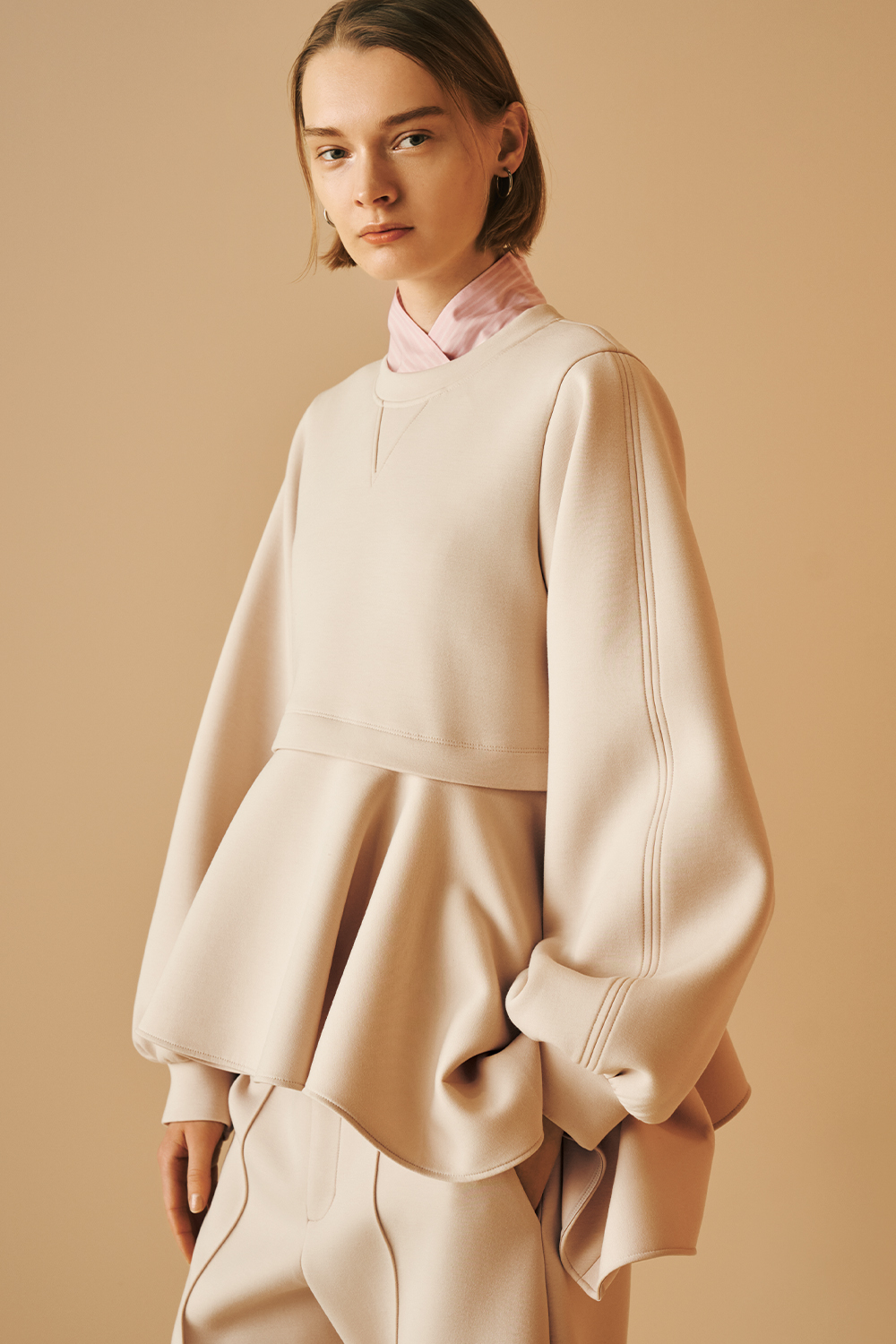 Model is wearing a beige oversized peplum top with voluminous sleeves, wide-leg matching pants.