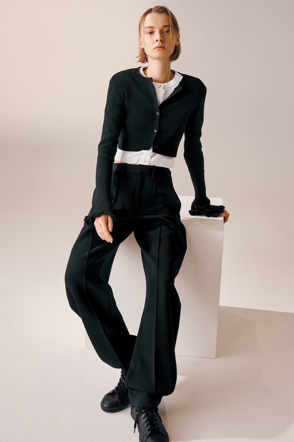 Model is wearing a cropped black cardigan over a white top, paired with high-waisted black flared trousers and black lace-up sneakers.