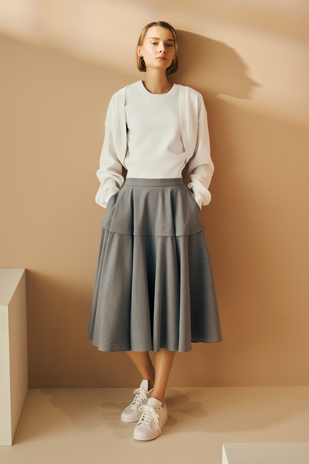 Model is wearing a white long-sleeve top with a layered detail, a gray midi flared skirt, and white lace-up sneakers.