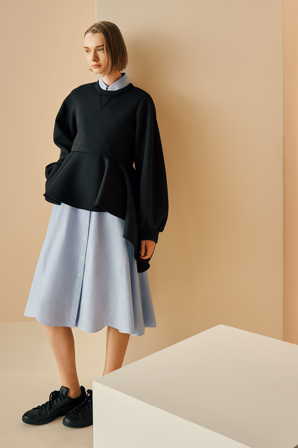 Model is wearing a black structured long-sleeve top layered over a light blue button-down shirt dress, paired with black lace-up sneakers.