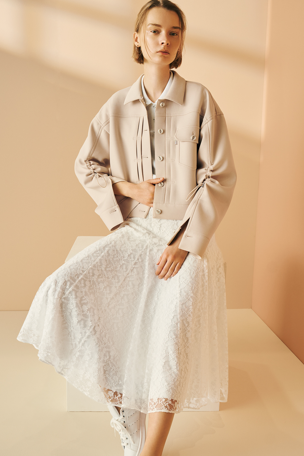 Model is wearing a beige oversized jacket over a light top, paired with a white lace midi skirt and white lace-up sneakers.