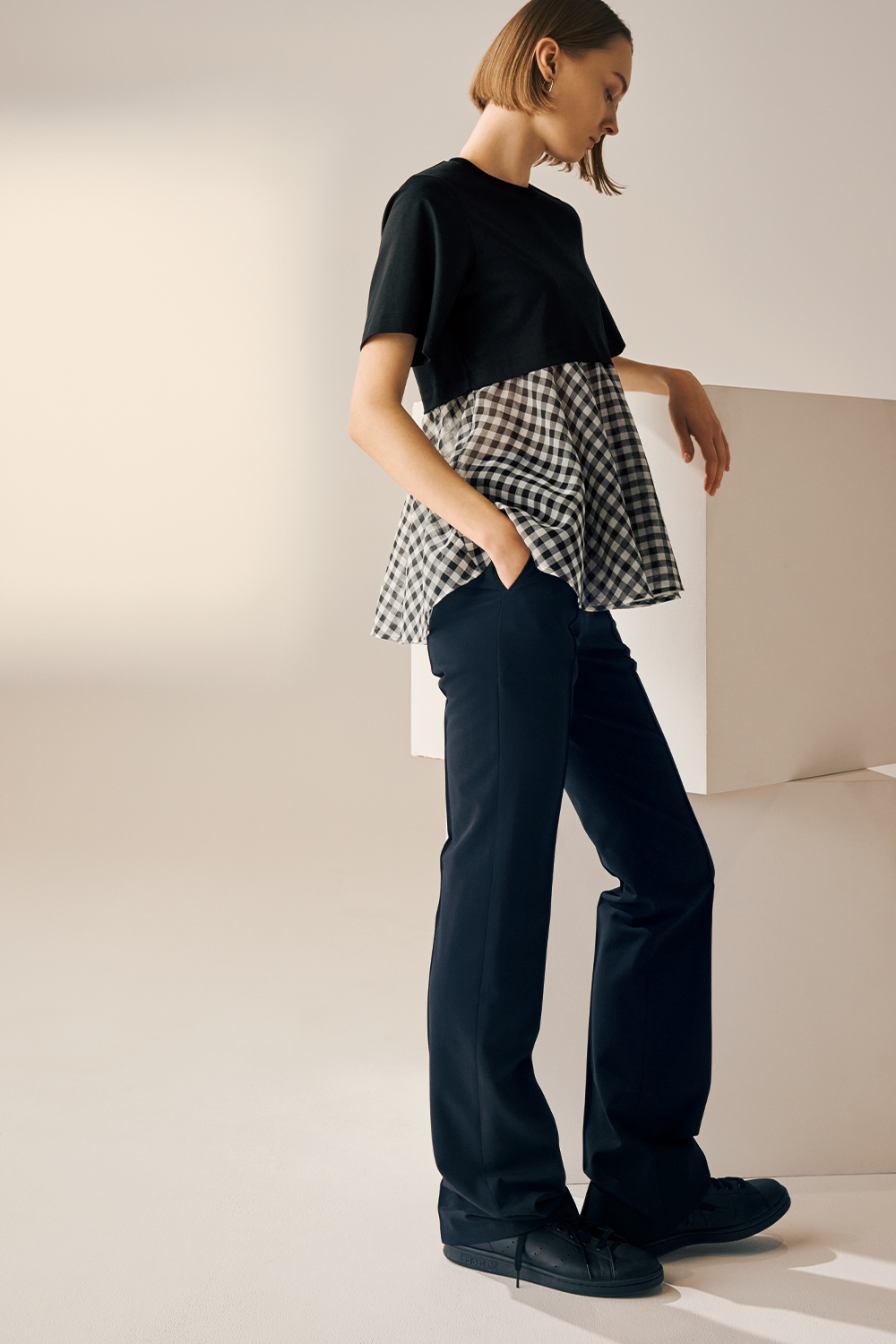Model is wearing a black short-sleeve top with a layered black-and-white gingham peplum hem, paired with navy wide-leg trousers and black sneakers.